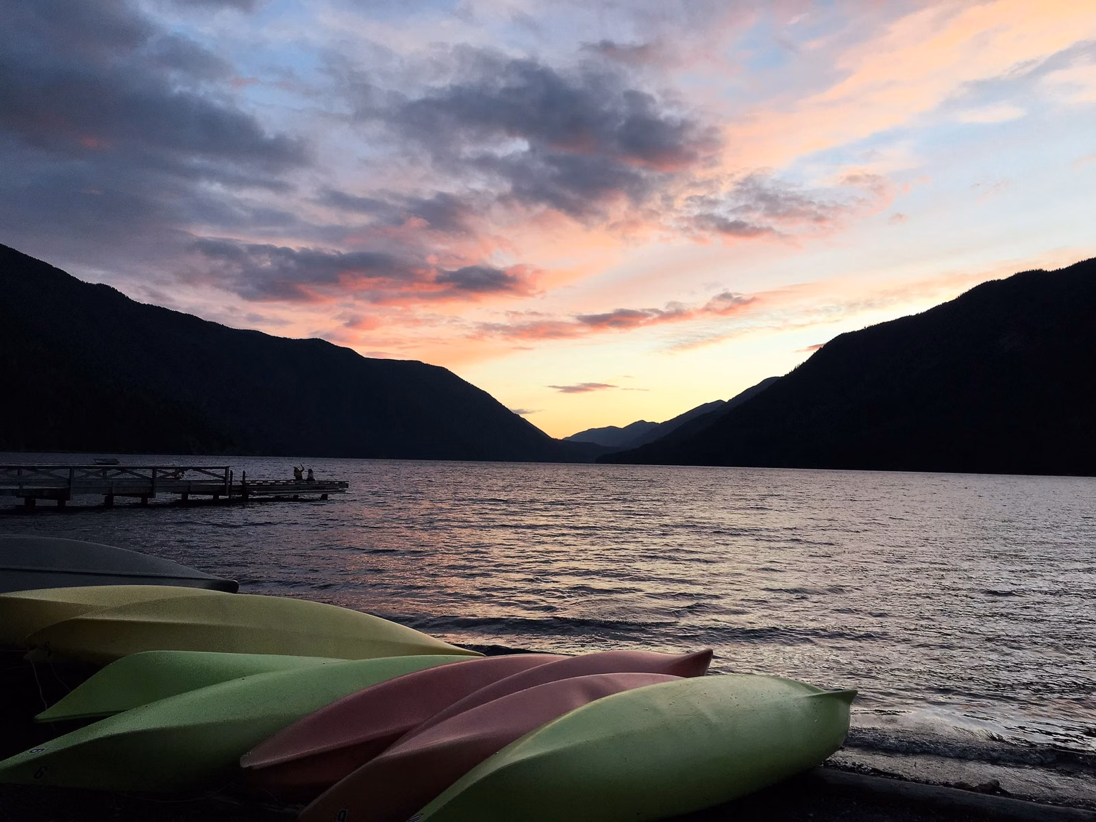 Kayaks pulled up on a lake beach at sunset with a dock and hills beyond