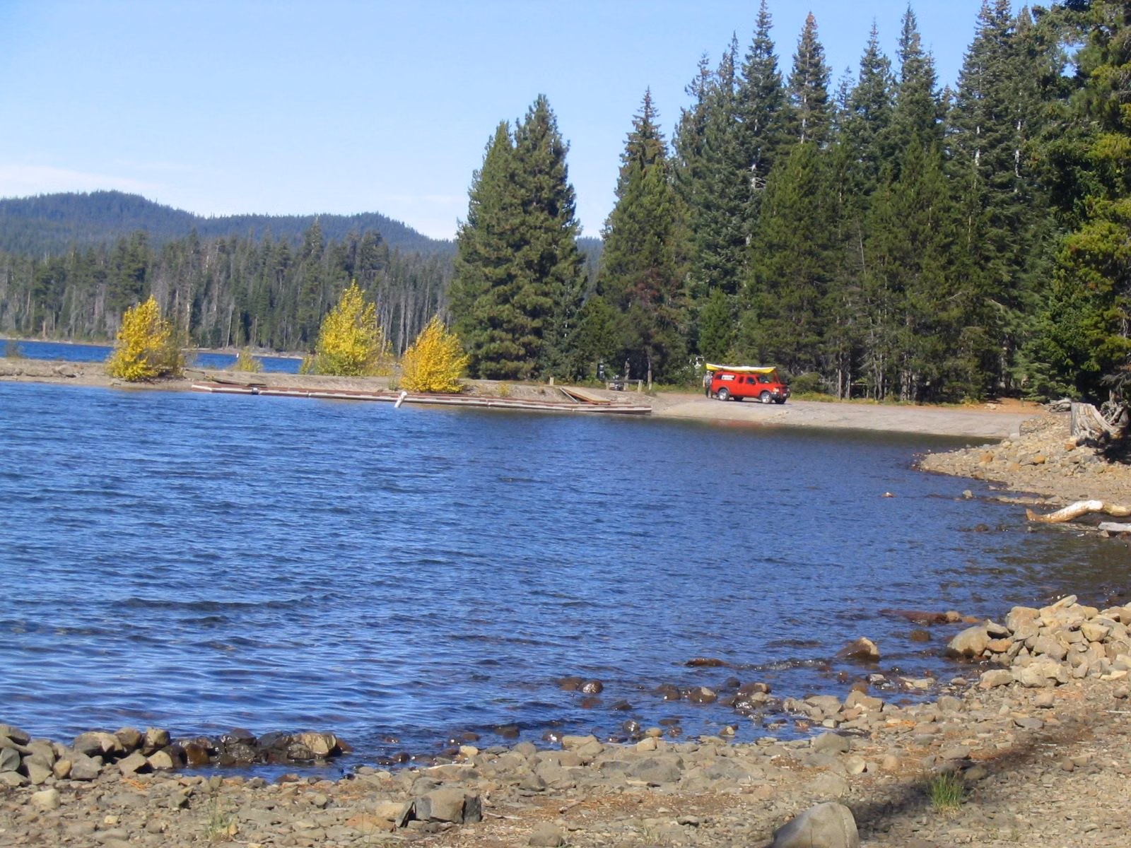 A car parked by a rocky lakeshore with firs and blue water