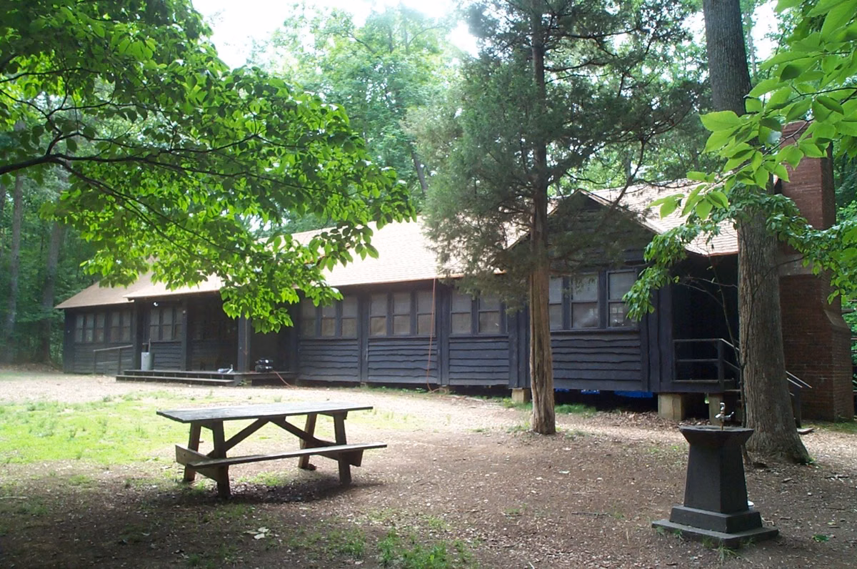 A long rustic dining hall with a picnic table in front, under shade trees